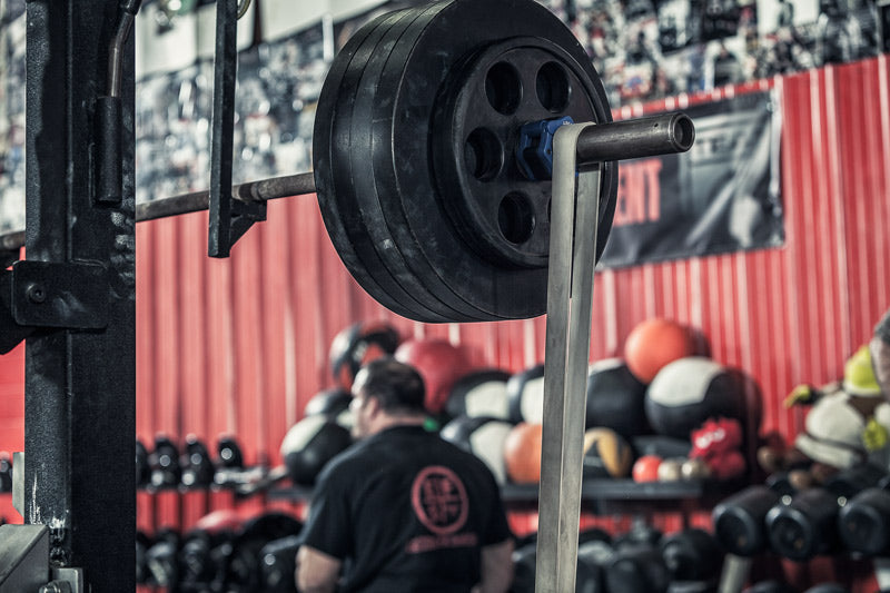 Barbell on a rack in a gym setting with various weights and equipment in the background.