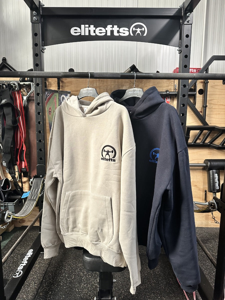 Two hoodies, one beige and one navy, hanging on a rack with 'elitefts' branding in a gym setting.